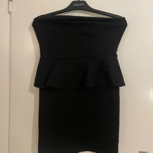 Svart axelbandslös peplumklänning - Säljer en svart, axelbandslös peplumklänning från KappAhl. Klänningen är kort, har en snygg volangdetalj i midjan och en guldig dragkedja bak. Sitter tight och har en stilren, elegant look som passar perfekt när du vill sticka ut.