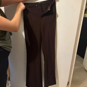 Bruna kostymbyxor från Gina tricot XS - Snygga bruna kostymbyxor från Ginatricot med klassisk rak passform och pressveck framtill. Byxorna har hög midja och stängs med knapp och dragkedja. Perfekta för en stilren look.