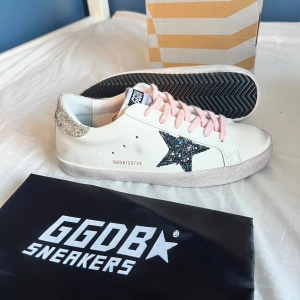 Golden Goose sneakers med glitterdetaljer - Snygga vita Golden Goose sneakers i skinn med rosa snören, glittrig blå stjärna på sidan och silverglittrig häl. Klassisk låg modell med platt sula och rund tå. Perfekta för dig som vill sticka ut med coola detaljer och en unik look. Storlek 39 men skulle säga att det passar 38 bättre. 