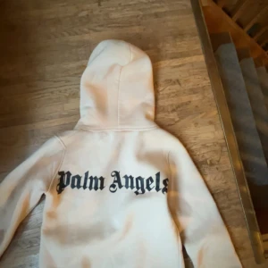 Vit hoodie från Palm Angels - Säljer en vit hoodie från Palm Angels med stor svart logga på ryggen och mindre logga framtill. Tröjan har huva med snörning och en klassisk känguruficka. Perfekt för dig som gillar streetwear och vill sticka ut med en snygg och clean look.