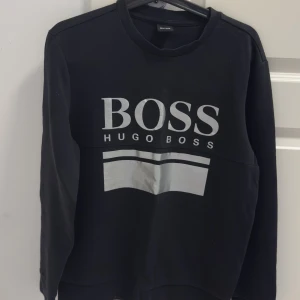 Svart sweatshirt från Hugo Boss - Säljer en svart sweatshirt från Hugo Boss med stor vit logga och grafiskt tryck framtill. Tröjan har rund halsringning och långa ärmar, perfekt för en stilren och avslappnad look.