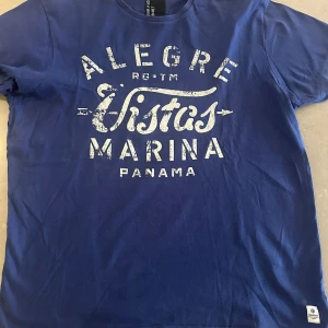 Blå t-shirt Fisher and Bennet M - Riktigt Grish Blå t-shirt från Fisher and Bennet i storlek M med vit, sliten tryckt text 'Alegre Vistas Marina Panama' på bröstet. Klassisk rund hals och korta ärmar. Skön och mjuk bomullskvalitet, perfekt för en avslappnad stil. Perfekt till sommarn för att vara lite unik