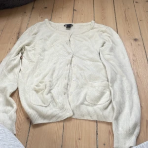 Beige kofta från H&M Basic, M - Beige kofta från H&M Basic i storlek M. Klassisk modell med rund halsringning, knappar framtill och två små fickor. Mjuk och skön, perfekt att slänga över en t-shirt eller linne. Ribbstickade muddar vid ärmslut och nederkant.