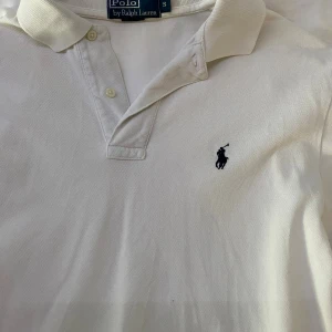 Vit pikétröja från Ralph Lauren - Klassisk vit pikétröja från Ralph Lauren med broderad logga på bröstet. Tröjan har krage, knäppning med två knappar och är kortärmad. Tillverkad i bomull för en skön och luftig känsla. Perfekt för en clean och stilren look.