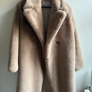 Beige pälsjacka från Dry Lake - Säljer en supermjuk och fluffig beige pälsjacka från Dry Lake. Jackan har bred krage, två knappar framtill och är lång i modellen. Perfekt för dig som vill ha en mysig och trendig look under kyliga dagar.