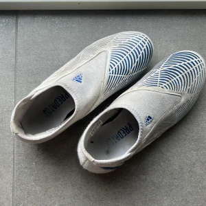 Adidas Predator fotbollsskor vita/blå - Säljer ett par Adidas Predator fotbollsskor i vitt med blåa detaljer och mönster. Skorna har en strömlinjeformad design utan snörning och är tillverkade i syntetmaterial. Sulan har fasta dobbar i vitt och blått för bra grepp på planen. Perfekta för dig som vill sticka ut på matchen.