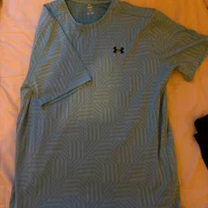 Ljusblå mönstrad t-shirt från Under Armour - Snygg ljusblå t-shirt från Under Armour med geometriskt mönster över hela tröjan. T-shirten har korta ärmar och en klassisk passform, perfekt för träning eller chill. Materialet är lätt och andas, vilket gör den skön att ha på sig under aktiva dagar.