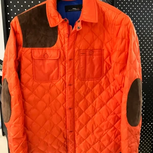 Quiltad orange jacka RLX Ralph Lauren - Quiltad orange jacka från RLX Ralph Lauren med bruna mockadetaljer på axel och armbågar. Två bröstfickor med knappstängning och klassisk krage. Jackan har en sportig streetvibe och är tillverkad i polyester med inslag av mocka. Nypris ca 3000-4000kr. 