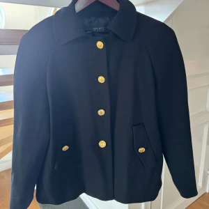 Svart kappa från Zara med guldknappar - Stilren svart kappa från Zara med klassisk krage och snygga guldknappar framtill. Jackan har två fickor med lock och en rak passform. Perfekt för dig som vill ha en tidlös och elegant look. Storlek M men passar en S