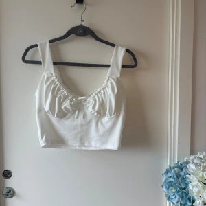 Vit croppad linnetopp med rosett - Säljer en vit croppad linnetopp med breda axelband och söt liten rosett framtill. Toppen har rynkade detaljer över bysten och är tillverkad i en mjuk bomulls- och polyesterblandning. Perfekt för en fräsch och stilren look.