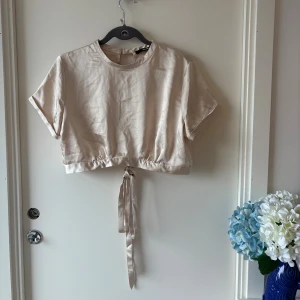 Champagnefärgad croppad topp Bik Bok - Snygg croppad topp i champagnefärgat satin från Bik Bok. Toppen har korta ärmar, rund halsringning och knytband i midjan. Baksidan är öppen med knapp i nacken och knyt längst ner för en extra cool detalj.