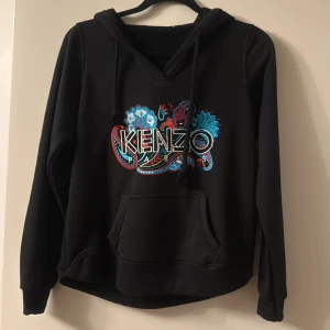 Svart hoodie med brodyr - Svart hoodie med färgglad broderad logga och blommönster på bröstet. Tröjan har huva med snörning, magficka och långa ärmar. Perfekt för dig som vill sticka ut med en snygg streetstyle-look.