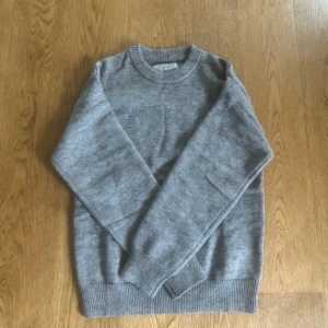 Grå stickad tröja med rund hals - En klassisk grå stickad tröja med rund halsringning och ribbade muddar vid ärmslut och nederkant. Aldrig använd.