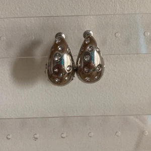 Silvriga örhängen med glitterstenar - Unika droppformade örhängen i silverfärg med glittrande stenar infällda över hela ytan. De har en chunky, rundad form som verkligen syns och ger en cool statement-look. Perfekta för dig som gillar att sticka ut med accessoarerna.