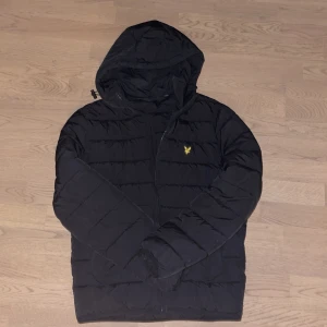 Svart pufferjacka från Lyle & Scott - Snygg svart pufferjacka från Lyle & Scott med huva och dragkedja. Jackan har ett litet gult emblem på bröstet och är perfekt för kyligare dagar. Den är både stilren och praktisk, vilket gör den till ett måste i garderoben.