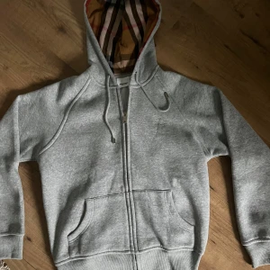 Grå hoodie från Burberry med dragkedja - Snygg grå hoodie från Burberry med klassiskt rutigt foder i huvan. Tröjan har dragkedja framtill, snörning i huvan och två fickor. Perfekt för dig som vill ha en stilren och lyxig vardagslook. Storlek xl men sitter som xs