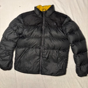 Svart pufferjacka från The North Face L - Svart pufferjacka från The North Face i storlek L. Jackan har gul insida, hög krage och dragkedja framtill. Klassisk quiltad design med två fickor på sidorna och diskret logga på bröstet. Perfekt för kalla dagar och riktigt snygg streetstil.