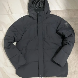 Svart pufferjacka från Five Seasons - Svart pufferjacka från Five Seasons med huva och diskret logga på ärmen. Jackan har en quiltad look, dragkedja framtill och innerfoder med camouflagemönster. Perfekt för kalla dagar och har en clean, modern vibe. Priset kan diskuteras skriv för fler bilder. Köpt för 2200 kr från Bozzt