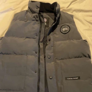 Grå dunväst från Canada Goose - Snygg grå dunväst från Canada Goose med hög krage, två stora fickor framtill och klassisk logotyp på bröstet. Västen har både dragkedja och tryckknappar för extra skydd mot kylan. Perfekt för lager-på-lager under kalla dagar.