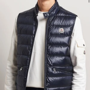 Mörkblå dunväst från Moncler - Snygg mörkblå dunväst från Moncler med quiltad design och hög krage. Västen har dragkedja framtill, två fickor med dragkedja och Moncler-logga på bröstet. Perfekt för lager-på-lager och stilren look. FINNS I FLERA STORLEKAR 