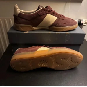 Vinröda sneakers från BOSS i mocka - Snygga vinröda sneakers från BOSS med beige detaljer och gummisula. Ovandelen är i mocka och skinn, med klassisk snörning och låg profil. Perfekta för dig som gillar stilrena och sportiga skor med premiumkänsla.