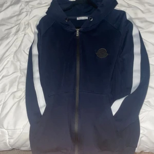 Mörkblå Moncler hoodie med vita ränder - Mörkblå hoodie från Moncler med vita ränder längs ärmarna och klassisk logga på bröstet. Tröjan har dragkedja framtill, huva och två fickor. Perfekt för dig som gillar sportig och stilren streetwear.