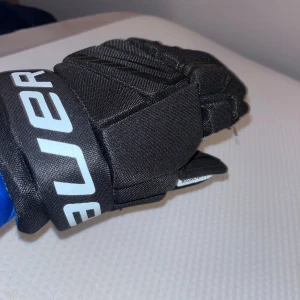 Svarta hockeyhandskar Bauer 10 tum - Säljer ett par svarta hockeyhandskar från Bauer i storlek 10 tum. Handskarna har blå detaljer på insidan av fingrarna och ett coolt mönster på fodret. De är vadderade och har vit Bauer-logga på mudden. Perfekta för dig som lirar hockey och vill ha bra skydd. Nypris 699 SEK