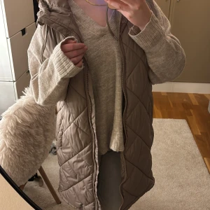 Lång quiltad dunväst med huva - Snygg lång dunväst i beige med quiltat mönster och dragkedja framtill. Västen har en huva med snörning och är perfekt att slänga över en stickad tröja. Stilren och enkel design som passar till många outfits.