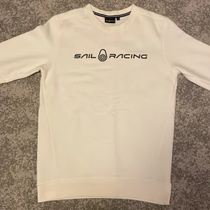 Vit Sail Racing sweatshirt i S - Vit sweatshirt från Sail Racing med svart logga framtill och en liten logga i nacken. Klassisk rund halsringning och långa ärmar. Tröjan har en clean och sportig look, perfekt för dig som gillar stilrena plagg. Materialet känns mjukt och bekvämt.