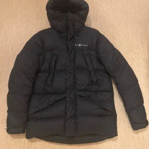 Svart dunjacka från Sail Racing - Svart dunjacka från Sail Racing med hög krage och huva. Jackan är fylld med certifierat dun (700 FP) och har flera praktiska fickor samt dragkedja framtill. Materialet är vindtätt och andas, perfekt för kalla vinterdagar. Snygg och funktionell design med diskret logga på bröstet.