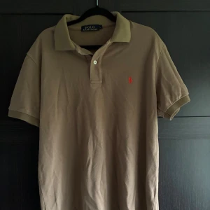 Beige pikétröja från Polo Ralph Lauren - Passar m och l Fin  pikétröja från Polo Ralph Lauren med röd broderad logga på bröstet. Tröjan har korta ärmar, krage och knäppning med två knappar. Perfekt för en avslappnad men ändå stilren look.