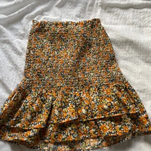 Blommig volangkjol från ONLY XS - Supersöt kort kjol från ONLY i storlek XS med blommigt mönster i orange, gul, svart och vit. Den kan även användas som en söt topp.Kjolen har smockad midja och volang nertill som ger en lekfull vibe. Perfekt för dig som gillar färg och mönster!