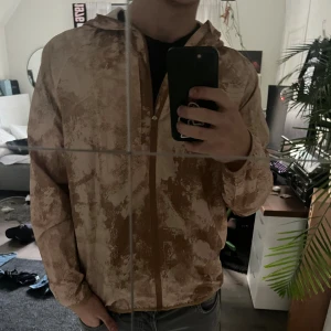 Beige och brun vindjacka med huva - Säljer en beige och brun vindjacka med camo-inspirerat mönster. Jackan har huva, lång ärm och dragkedja framtill. Lätt och smidig modell som passar perfekt till vår och höst.