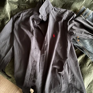 Mörkblå skjorta från Ralph Lauren - Snygg mörkblå skjorta från Ralph Lauren med klassisk krage och knappar hela vägen. Ikoniska röda loggan broderad på bröstet. Skjortan är långärmad och tillverkad i mjuk bomull, perfekt för en clean och stilren look.