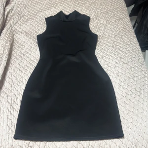 Svart kort klänning från Sisters Point - Snygg svart kort klänning från Sisters Point i figurnära modell. Ärmlös design med hög hals och en cool öppning i ryggen. Perfekt för dig som gillar stilrena och trendiga plagg. Materialet känns mjukt och stretchigt.