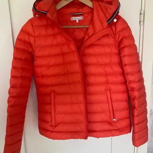 Orange/ röd pufferjacka från Tommy Hilfiger - Säljer en orange/ röd  snygg pufferjacka från Tommy Hilfiger i storlek S. Jackan har huva, dragkedja framtill och två fickor med dragkedja. Perfekt för kyliga dagar och har en klassisk quiltad look. Passar dig som gillar sportig och stilren vibe.