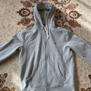 Grå zip Ralph Lauren - Grå hoodie från Polo Ralph Lauren med dragkedja framtill, två fickor och vit snörning i huvan. Klassisk broderad logga på bröstet.