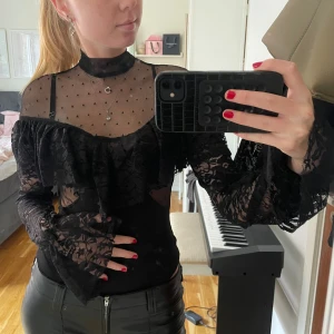 Svart spetsblus i body med volang  - Supersnygg svart blus med transparent mesh upptill och prickigt mönster. Långa ärmar i spets och volangdetalj över bysten och axlarna. Stilren med figurnära passform, perfekt för att sticka ut. Perfekt för fest eller middag!
