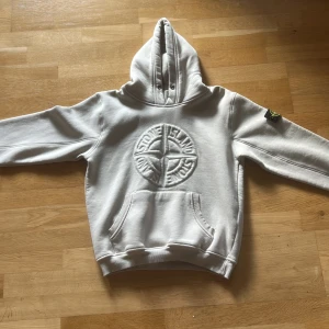 Vit hoodie från Stone Island - Säljer en vit hoodie från Stone Island med broderad logga på bröstet och klassisk patch på ärmen. Tröjan har huva med snörhål, känguruficka och ribbade muddar. Perfekt för dig som gillar streetwear och vill ha något stilrent men ändå fett.