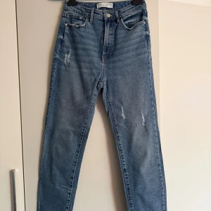 Stradivarius blåa mom jeans, storlek 38. Nyskick - •Snygga blå mom jeans från Stradivarius med hög midja och raka ben.  •Använda 2-3 gånger och tvättade. Som nya •Jeansen har slitna detaljer framtill, klassiska fem fickor och stängs med dragkedja och knapp.  •Perfekta för en avslappnad och trendig look. •Djurfritt och rökfritt hem.