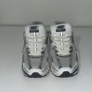 Nike Vomero 5 silver/vita sneakers - Nike Vomero 5 sneakers i silver och vitt med meshpaneler och reflekterande detaljer. Skorna har snörning, svart swoosh och en chunky sula. Perfekta för dig som gillar sportig och trendig stil. Snygga detaljer och bekväm passform.