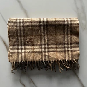Beige rutig halsduk från Burberrys - Klassisk halsduk från Burberrys i beige och bruna toner med rutigt mönster och fransar i ändarna. Broderad logga och tillverkad i 100% ull. Perfekt accessoar för att lyfta din outfit med en tidlös brittisk touch. En mindre modell än de vanliga Burberry halsdukarna
