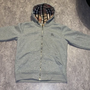 Grå Burberry hoodie med dragkedja - Grå hoodie från Burberry med klassiskt rutigt foder i huvan och insidan. Tröjan har dragkedja framtill, två fickor och broderad logga på bröstet. Perfekt för dig som vill ha en stilren och lyxig look med ikoniska Burberry-detaljer. Priset kan diskuteras. 