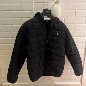 Svart pufferjacka från Lyle & Scott - Svart pufferjacka från Lyle & Scott med gul logga på bröstet. Jackan har quiltad design, dragkedja framtill och lång ärm. Perfekt för kyliga dagar och har en clean, sportig look. Bara att skriva om du har frågor!!