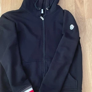 Svart Moncler hoodie med dragkedja - Svart hoodie från Moncler med hel dragkedja, huva med snörning och Moncler-logga på ärmen. Ribbade muddar med röd, vit och grå randig detalj. Tillverkad i mjuk bomullsmix, perfekt för höst och vinter. Klassisk och stilren design.