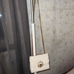 Guldig axelväska i tweed med kedja - Snygg axelväska i tweed med guldiga detaljer och kedjerem. Väskan har ett elegant rutigt mönster, ett stilrent spänne framtill och röd insida med dragkedjefack. Perfekt för dig som vill ha en chic och trendig accessoar.