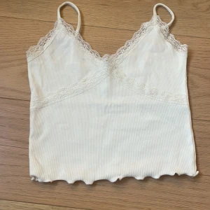 Vit spetslinnetopp från Gina Tricot - Supersöt vit linnetopp från Gina Tricot med tunna axelband och spetsdetaljer längs urringningen och under bysten. Toppen är ribbad och har vågig kant nertill, perfekt att styla med jeans eller kjol.
