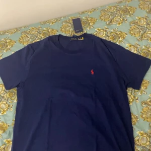 Marinblå t-shirt från Polo Ralph Lauren - Snygg marinblå t-shirt från Polo Ralph Lauren med klassisk röd logga broderad på bröstet. T-shirten har rund halsringning och korta ärmar. Perfekt basic-plagg med stilren design och skön passform.