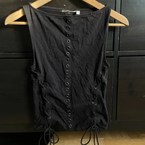 Svart korsett-topp med snörning från weekday - Svart korsett-inspirerad topp från weekday med coola metallöljetter längs hela framsidan och snörning på båda sidor. Ärmlös och tight passform, perfekt för att skapa en edgy look. Materialet är mjuk bomull som sitter skönt mot kroppen.