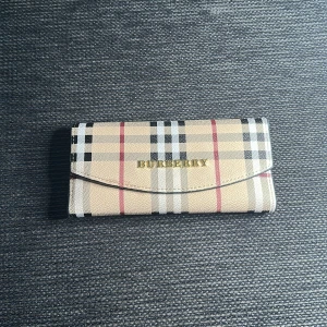 Rutig plånbok från Burberry - Snygg plånbok från Burberry i klassiskt beige, svart, vit och rött rutigt mönster. Plånboken har guldfärgad logga, tryckknapp och flera fack för kort och ID. Utsidan är i tåligt material och insidan är svart med stilren design.